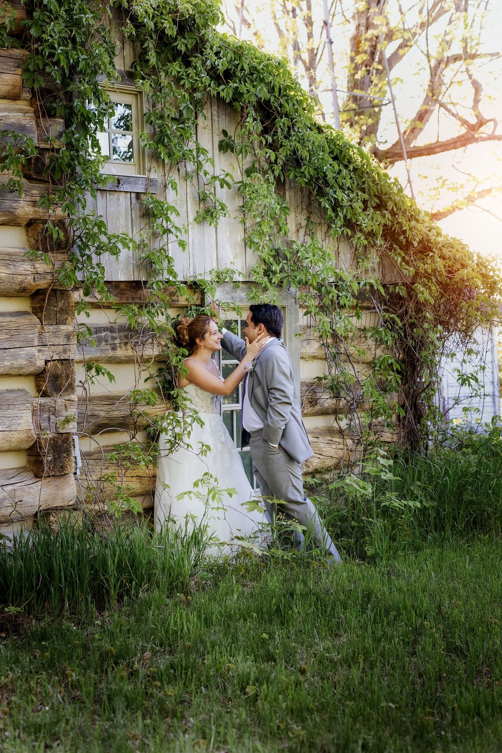 Bride and groom share a romantic moment by a rustic log cabin covered in lush green ivy on their wedding day.