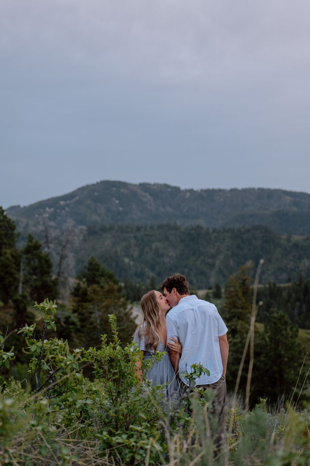 A romantic couple shares an intimate moment against a backdrop of mountain vistas and evergreen trees.