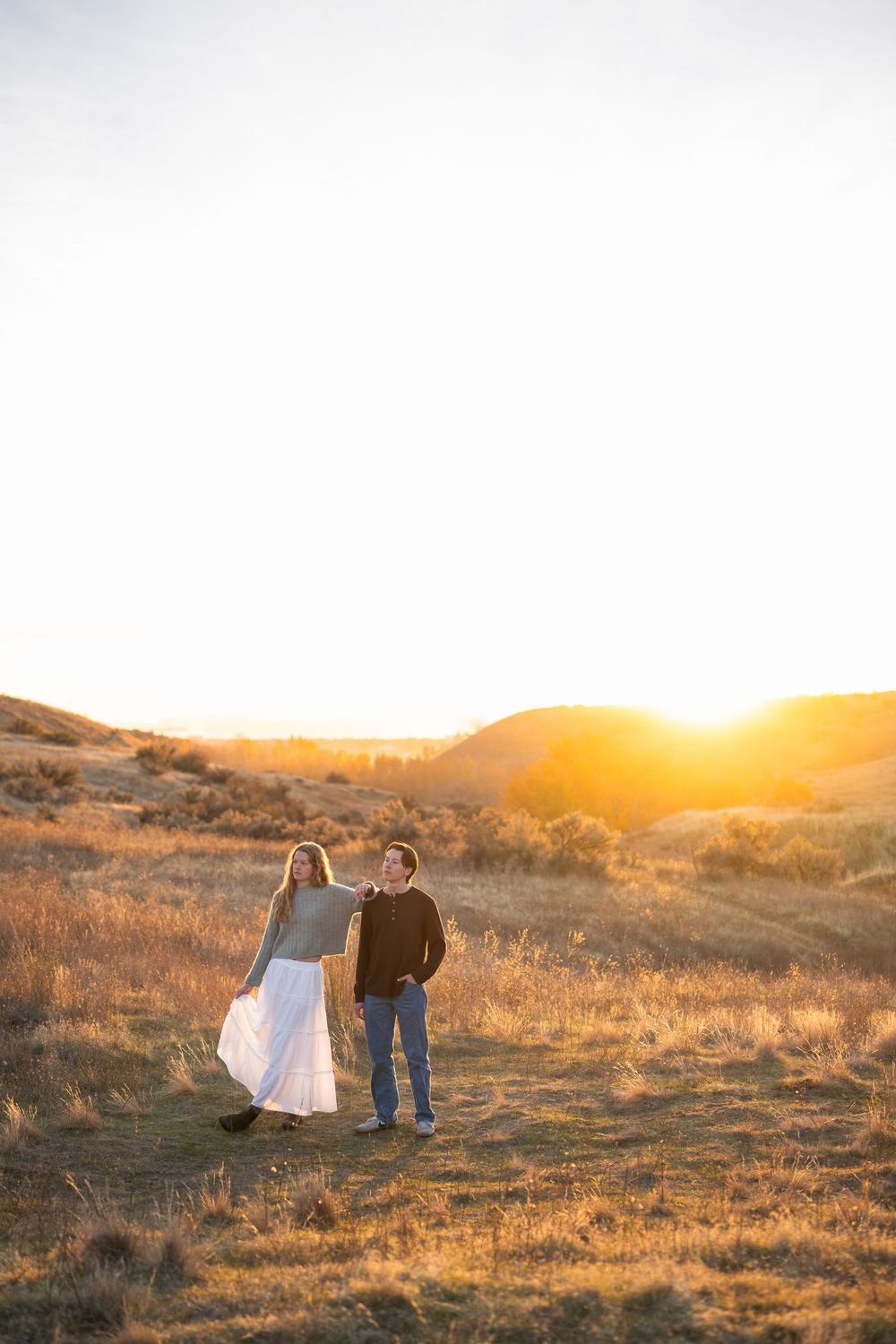 A couple stands together on a hillside during golden hour sunset in a desert landscape.