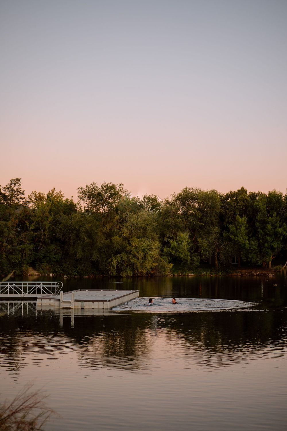 A tranquil lake scene at sunset with a wooden dock extending over calm waters surrounded by trees.