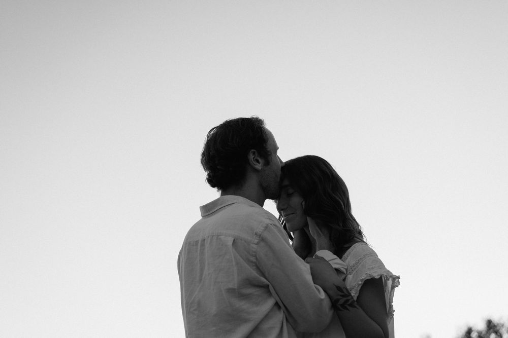 A black and white silhouette of a romantic couple embracing while standing against a bright sky.