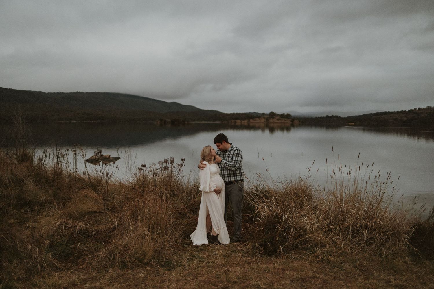 A pregnant woman and her partner embrace as they stand together next Googong dam water under gloomy skies at sunset.