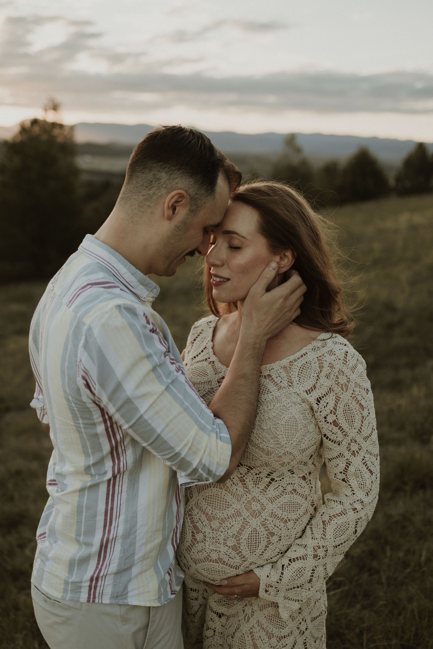 Romantic pregnant couple embraces in a lace dress and striped shirt against a sunset Canberra mountain landscape background.