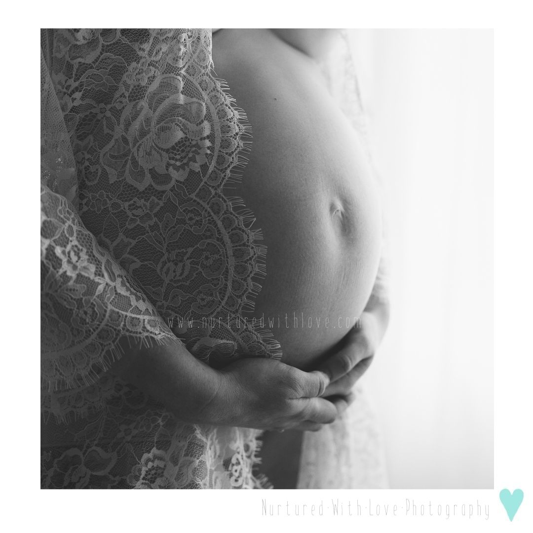 A delicate black and white maternity photo showing a pregnant belly draped in elegant lace against a light background.