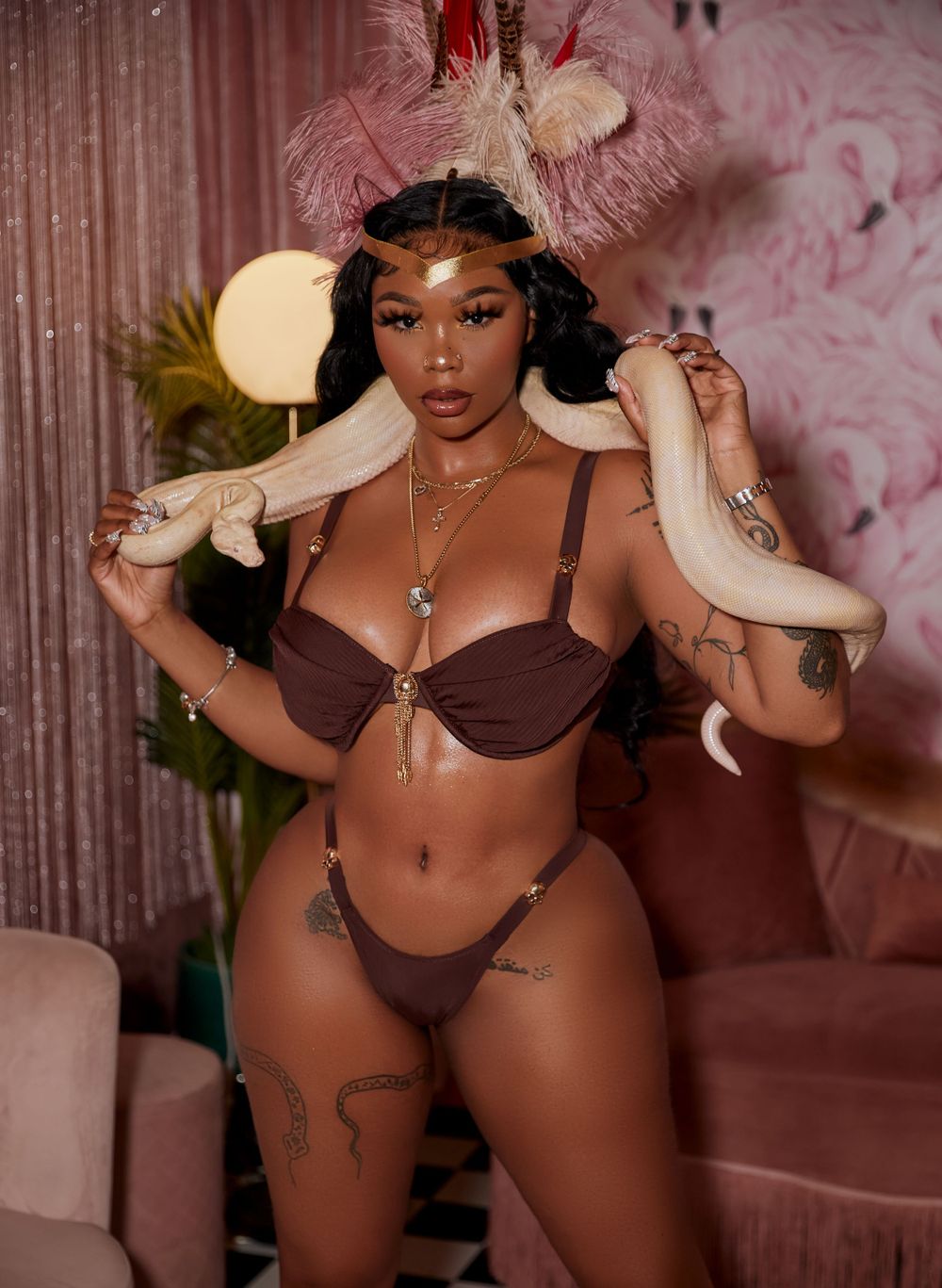 Model posing in brown lingerie with feather decorations in pink studio setting.