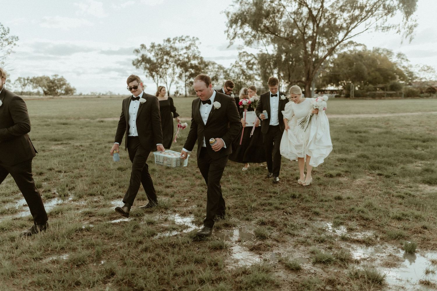 A group in formal attire walks across a grassy field on a cloudy day in a vintage-style photo.