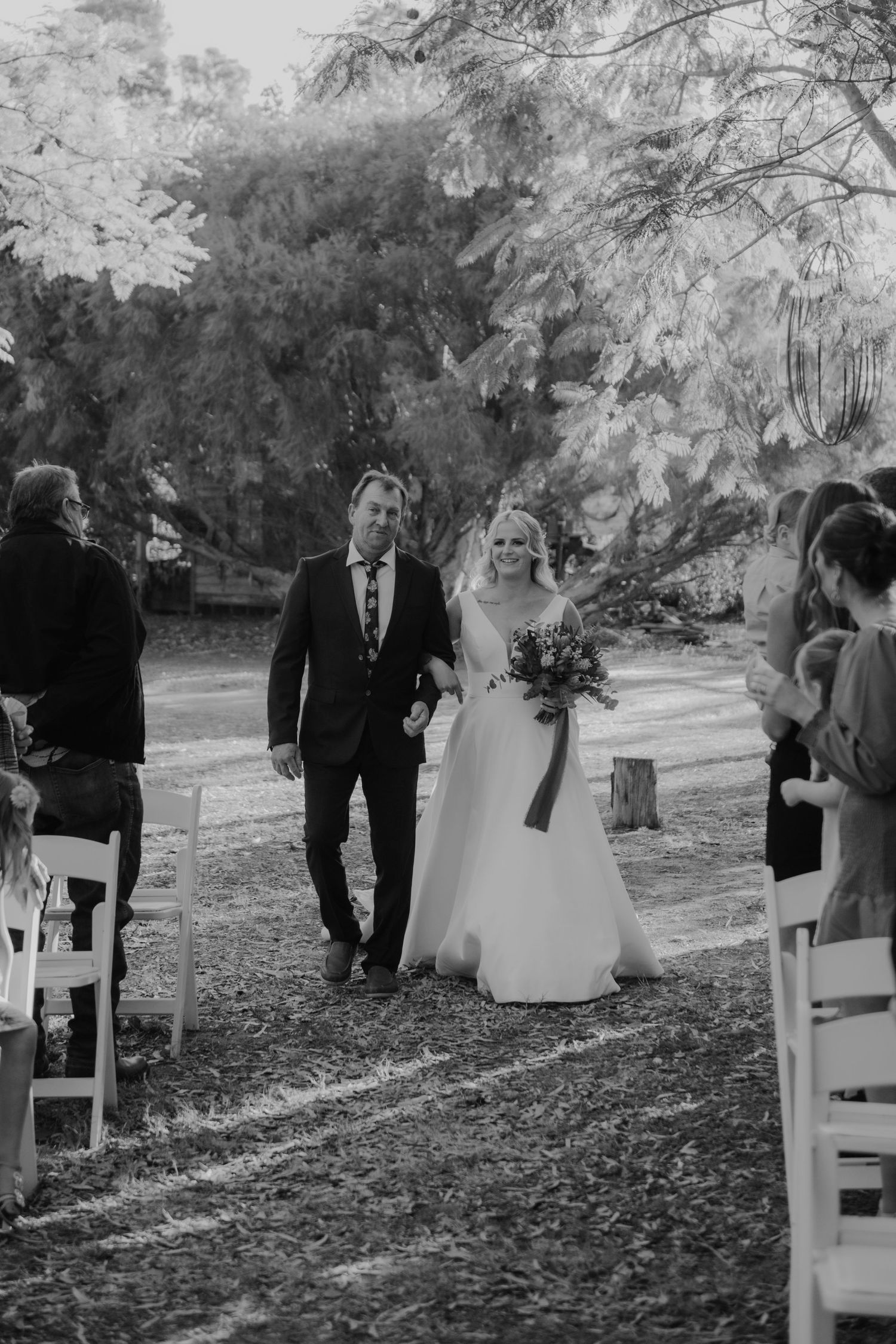 A black and white outdoor wedding ceremony scene with guests seated in white chairs along a tree-lined path.