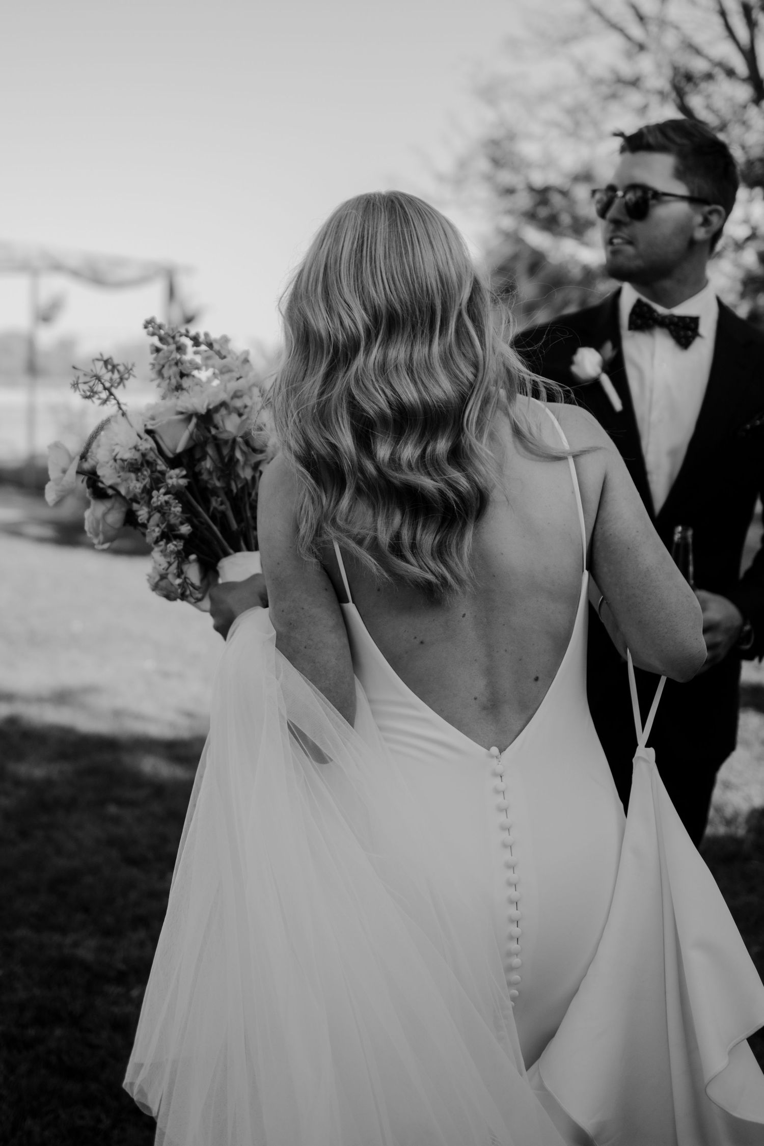 Black and white photo of a bride in backless dress with flowing hair holding bouquet next to groom in tuxedo outdoors.