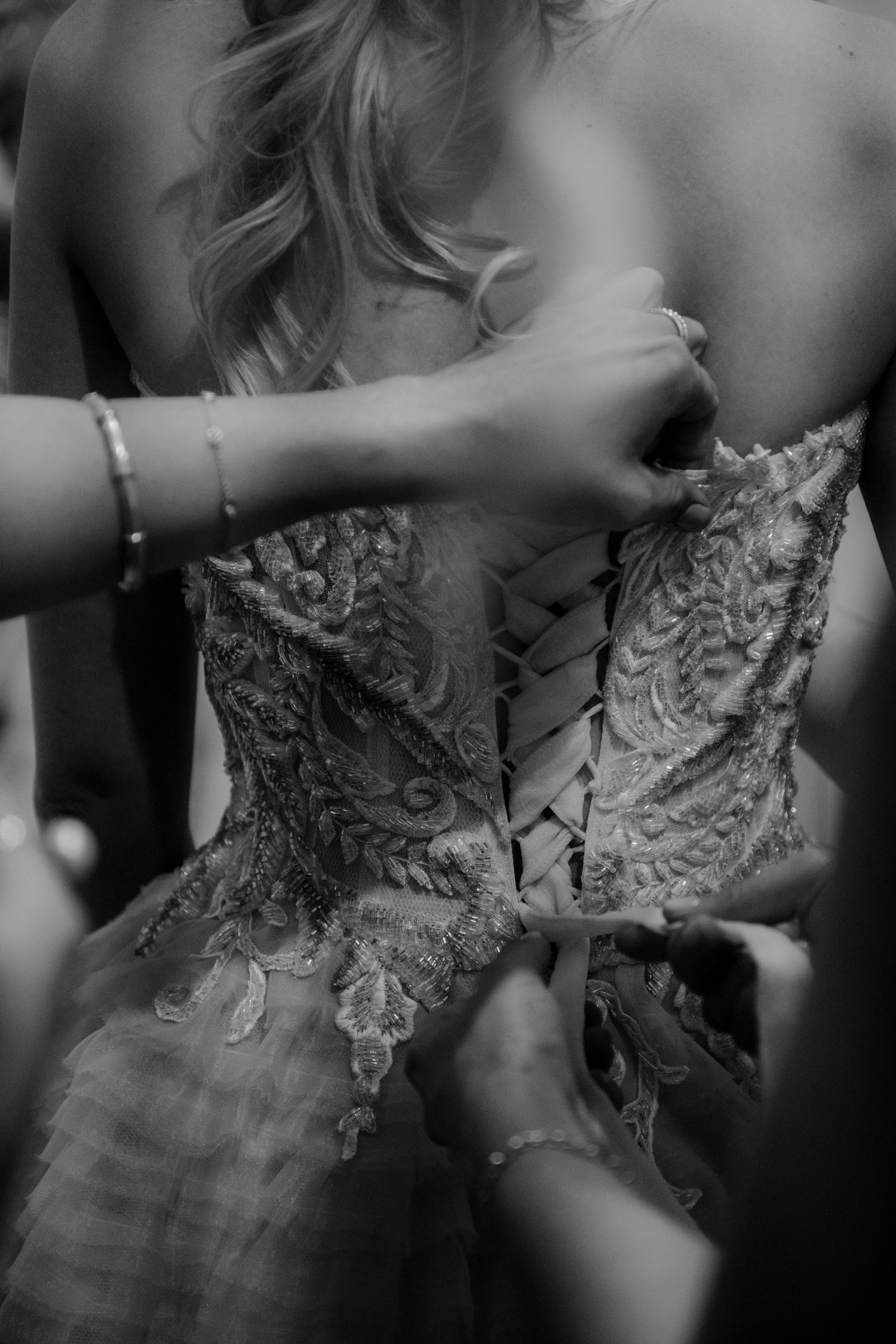 Hands lacing up an ornate wedding dress with delicate lace detail and corset-style back in black and white.