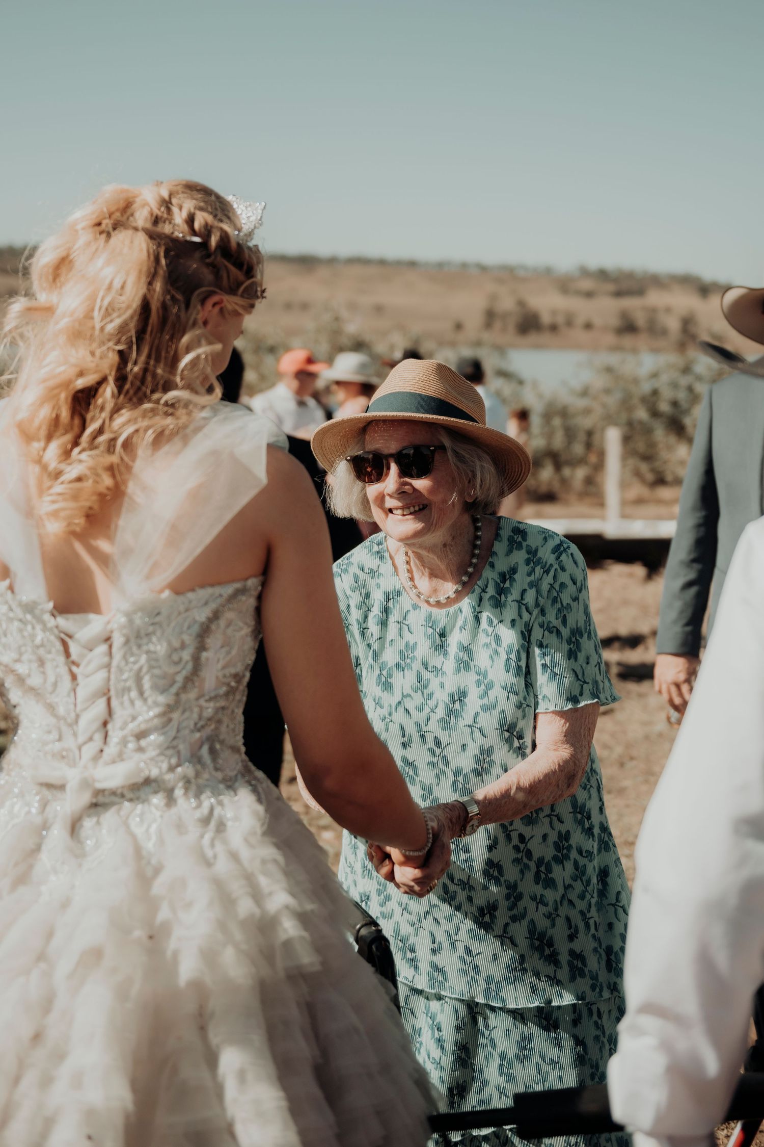 A bride in a white lace dress greets an elderly guest wearing a mint green floral dress and sun hat at an outdoor wedding.