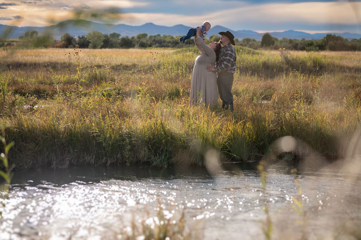 Family standing together in a golden meadow near a reflective stream with mountains behind.