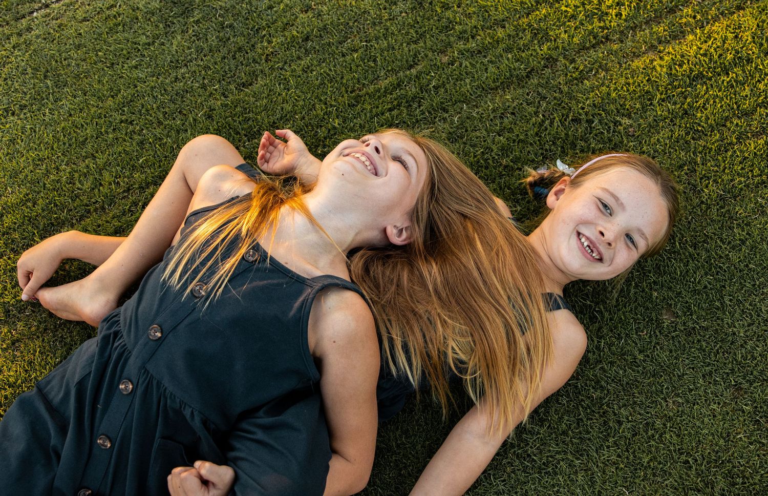 Two girls in black dresses run barefoot across vibrant green grass at golden sunset.