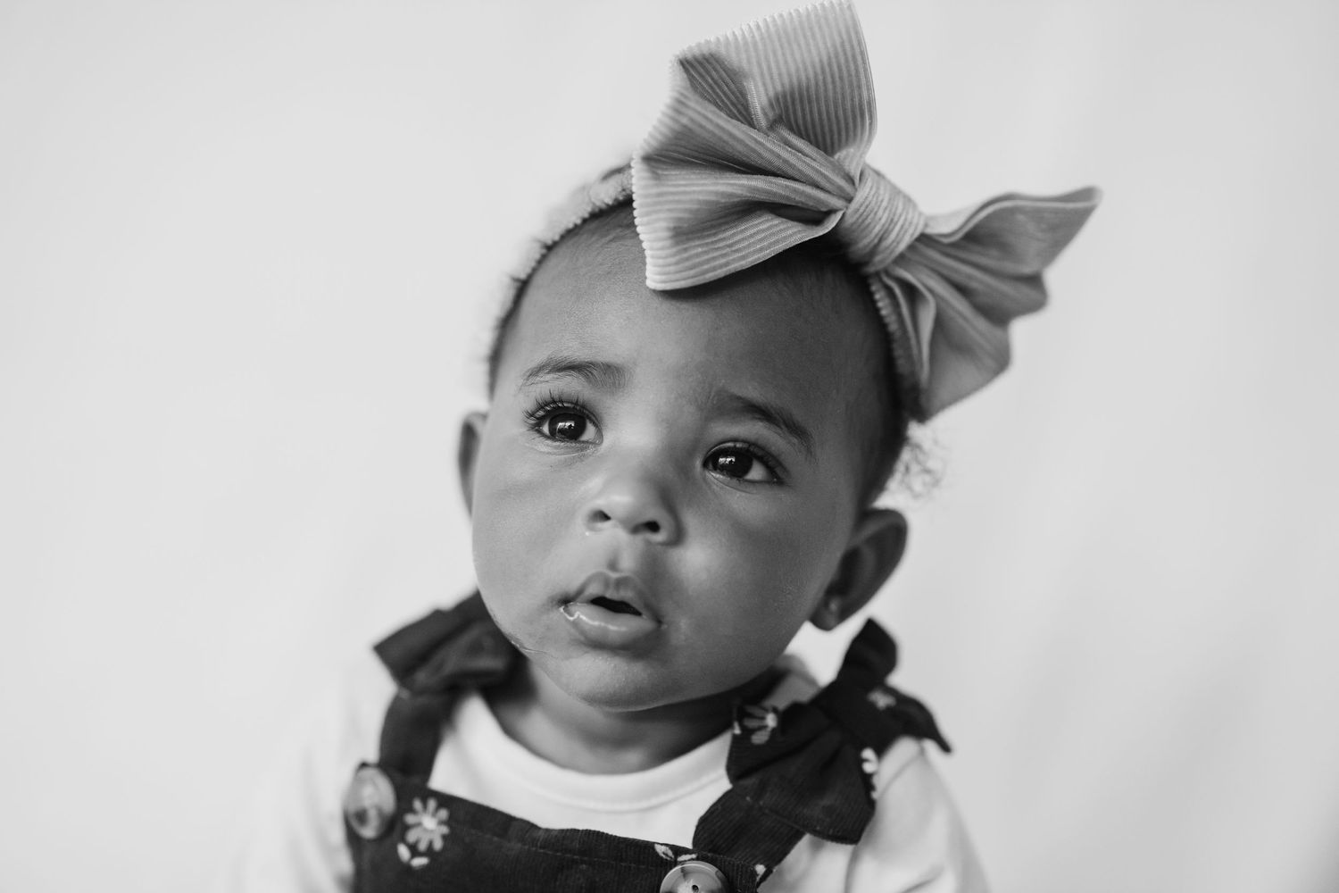 Black and white portrait of a baby wearing a large bow and overalls.