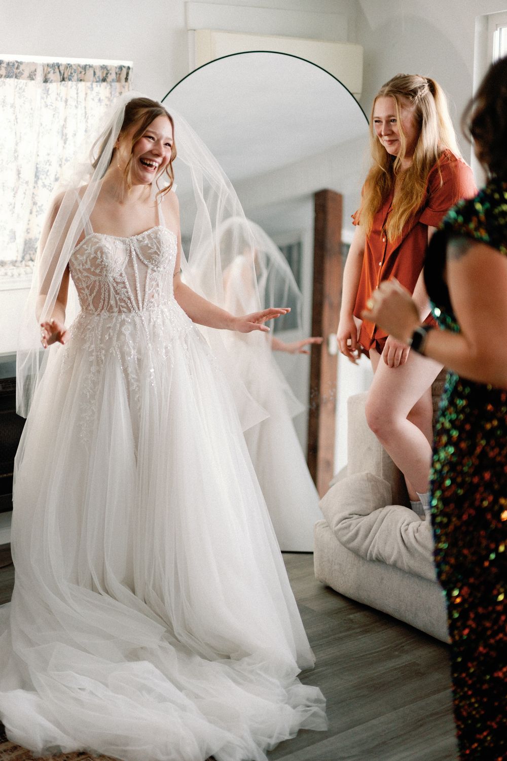 Bride laughing in a white tulle wedding gown and veil, surrounded by smiling bridesmaids in a bright dressing room.