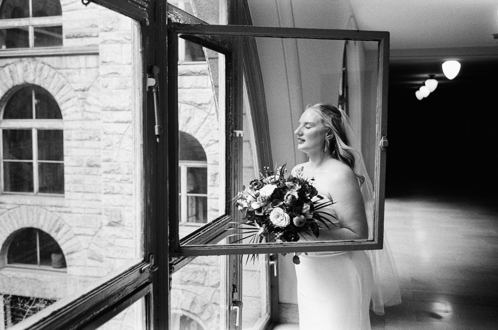 Bride in white gown holding bouquet reflected in open window, elegant black and white wedding photography.