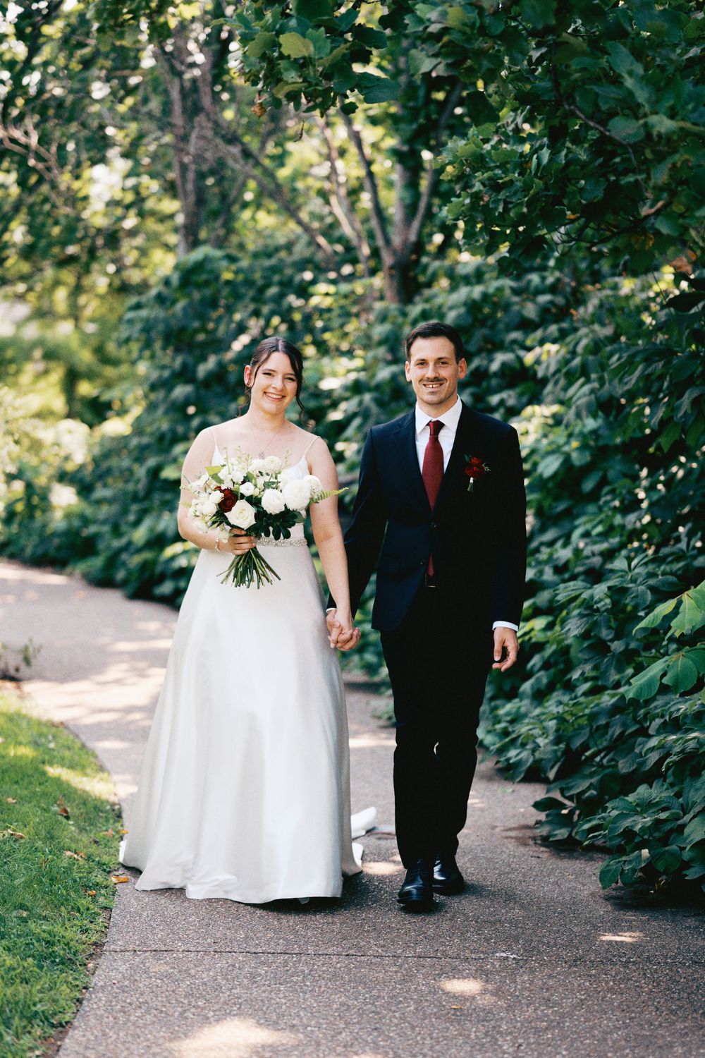 A bride and groom walk hand-in-hand on a garden path, she in a white gown with bouquet, he in a navy suit with red tie.