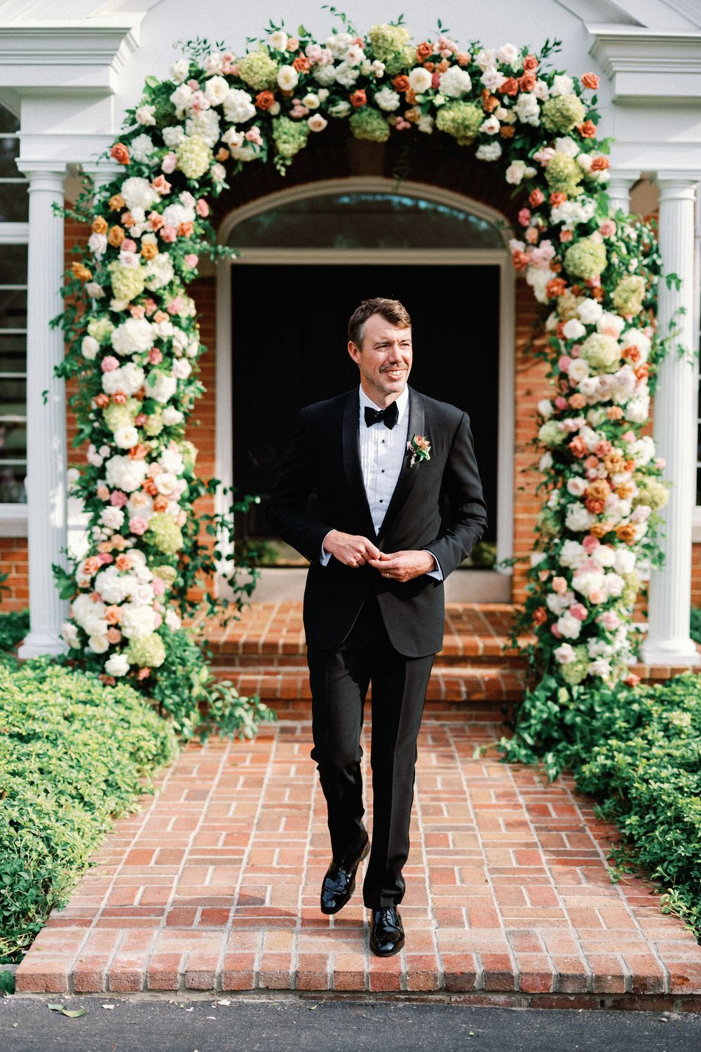 Classic black tuxedo and bowtie groom poses under beautiful peach and white floral wedding arch on brick steps.