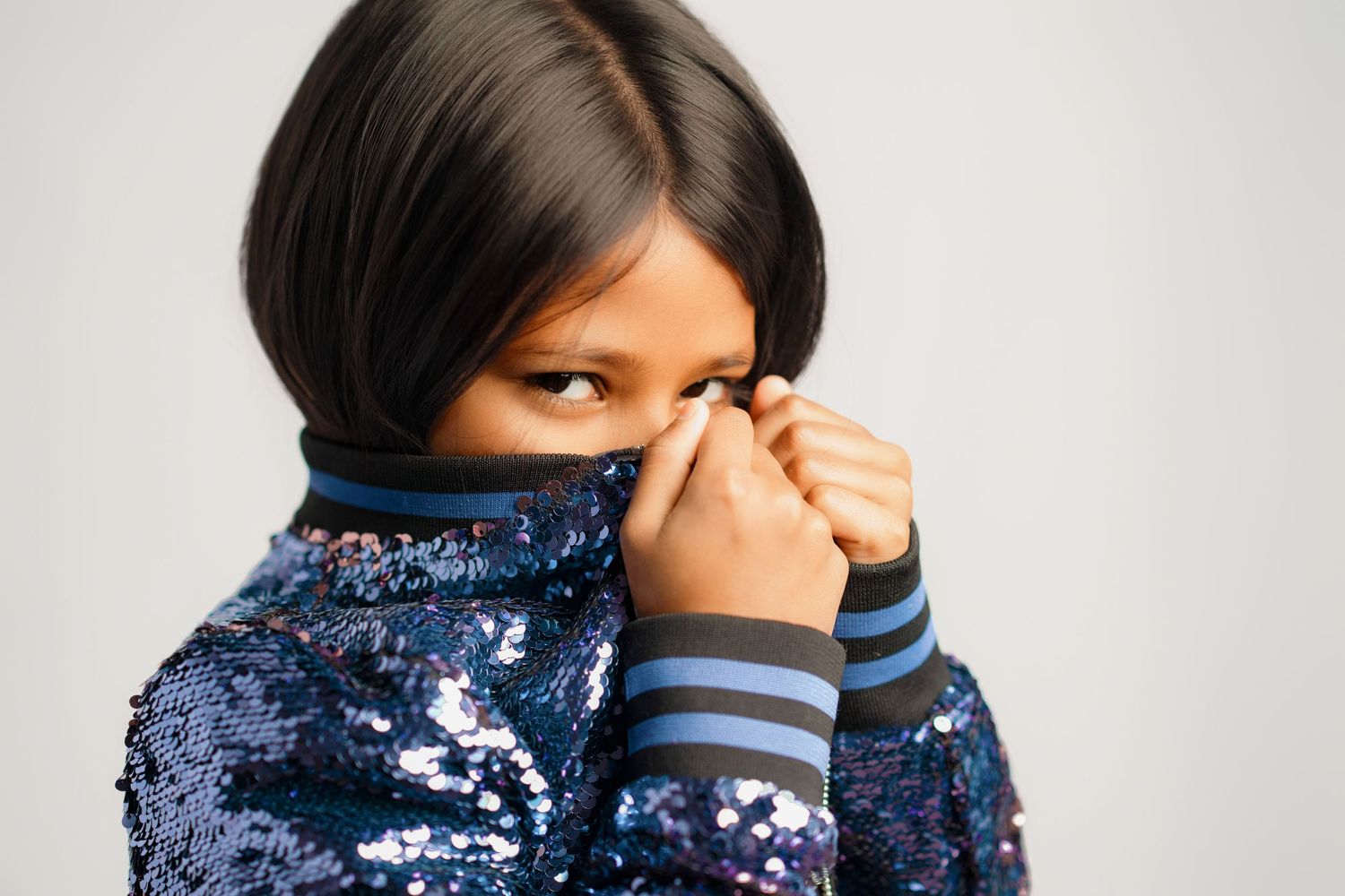 A person in a sparkly blue sequined jacket holds their sleeve up near their face while looking camera.
