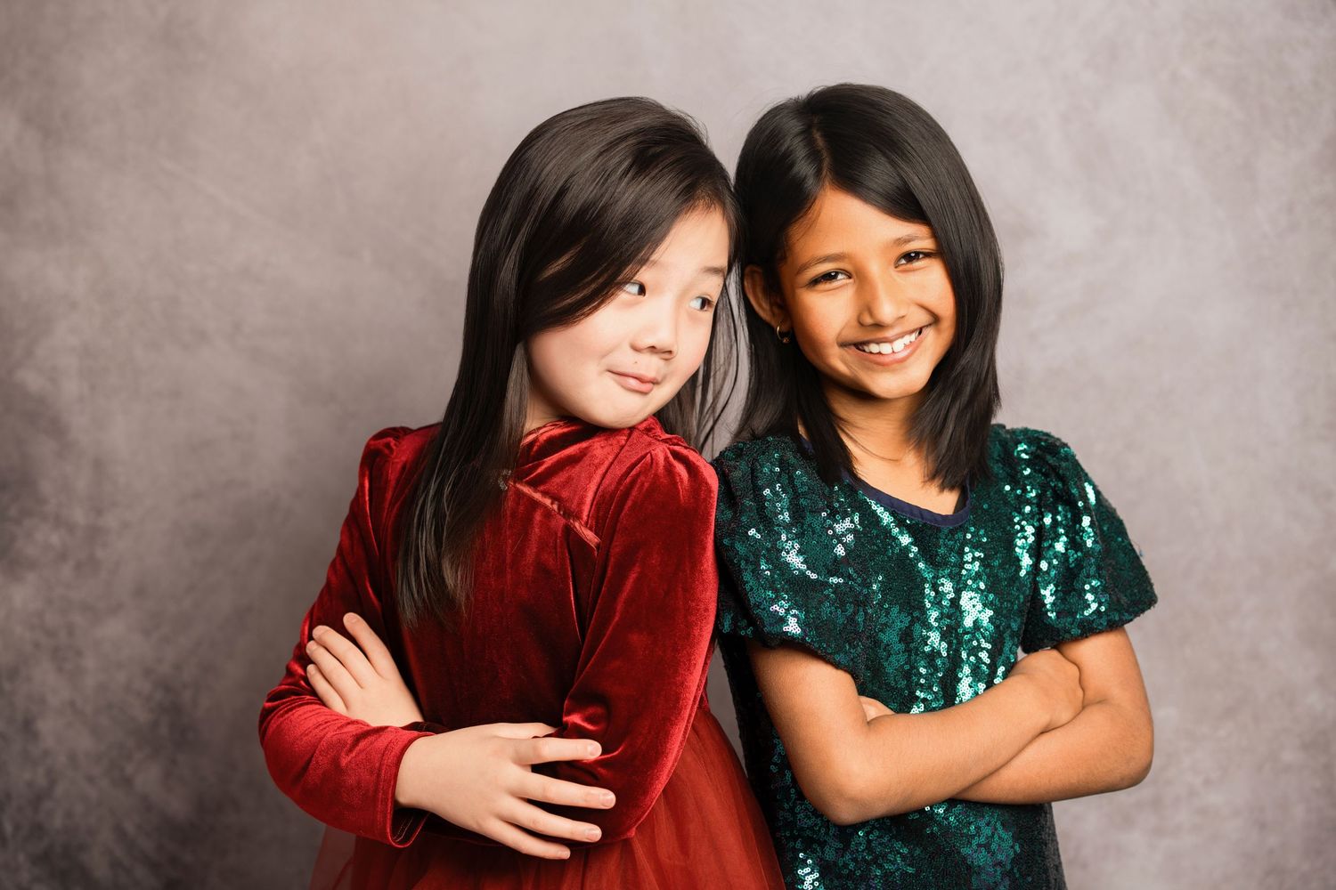 Two young girls pose with crossed arms wearing velvet holiday dresses in red and green sequins against a gray backdrop.