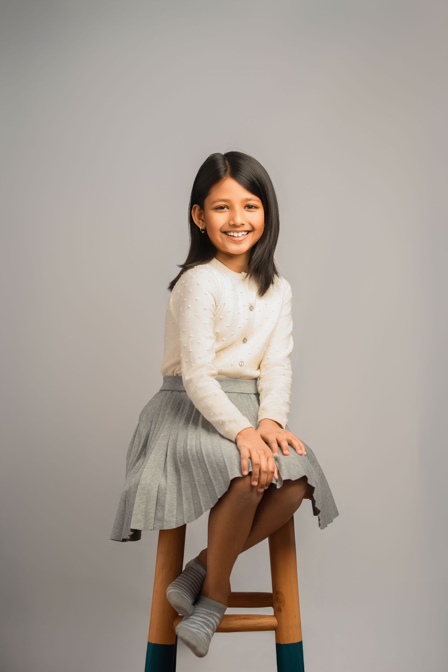 A young girl in a white sweater and gray pleated skirt sits on a wooden stool against a neutral background.