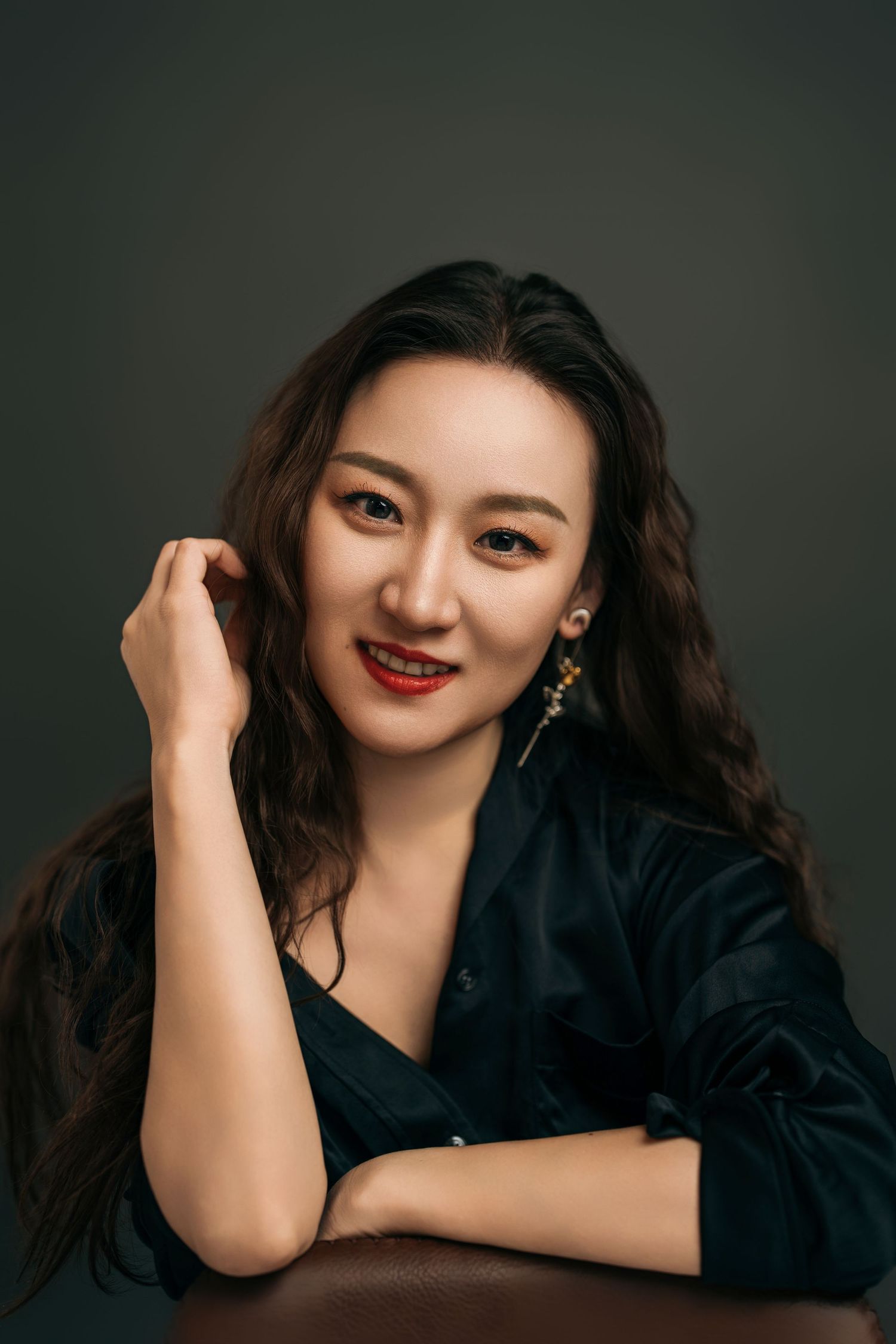 Professional portrait of someone wearing a black outfit and red lipstick against a dark background, with long wavy dark hair.