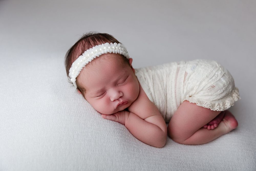 A newborn baby sleeping peacefully on a white backdrop wearing a headband and wrapped in soft fabric.