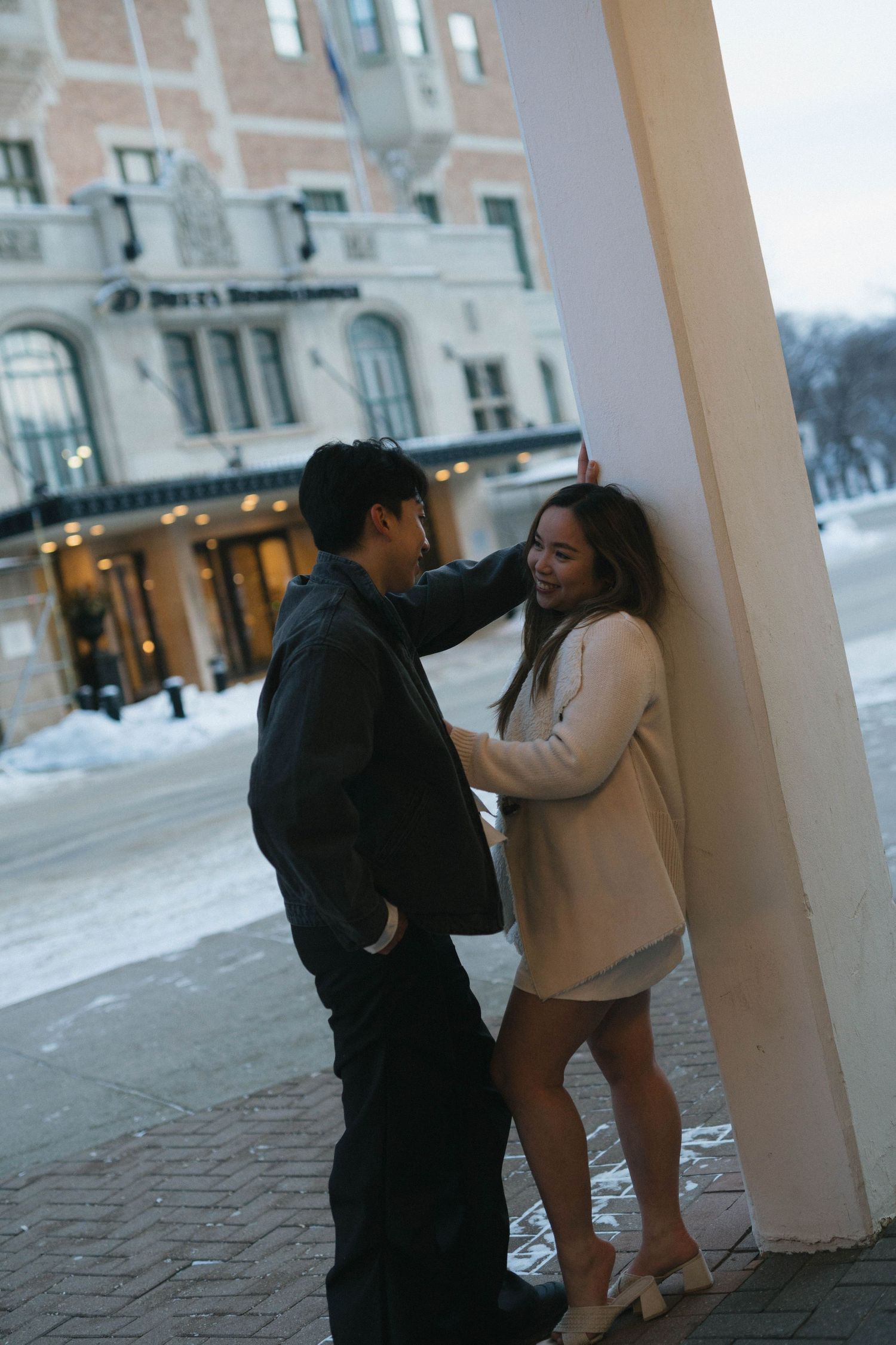 A couple shares an intimate moment under a column on a snowy winter evening in front of a lit building. Saskatoon Wedding Photographer