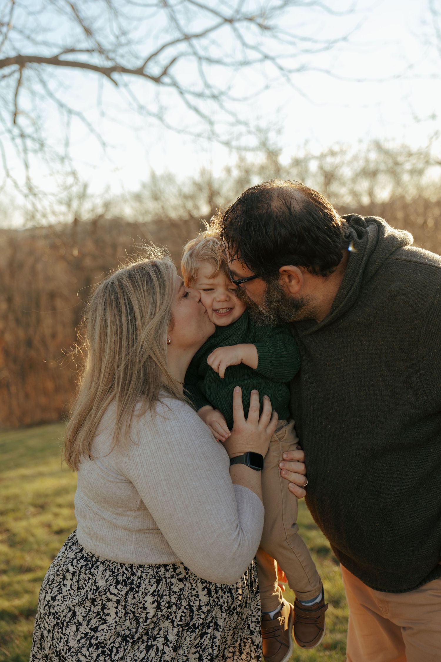 A tender family moment captured outdoors with warm sunlight filtering through bare branches.