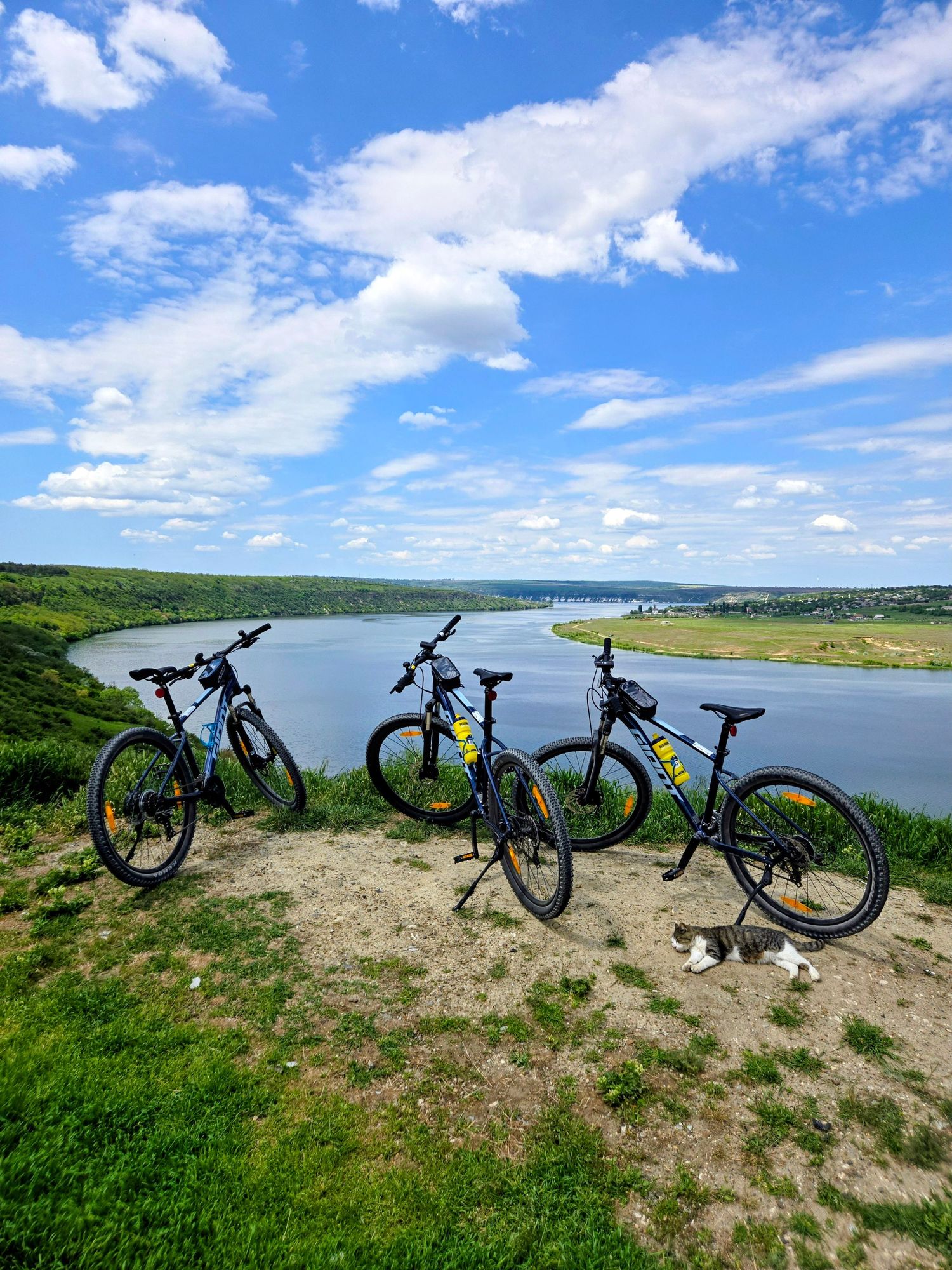 Three mountain bikes parked on a hilltop overlooking a serene blue river under a bright cloudy sky.