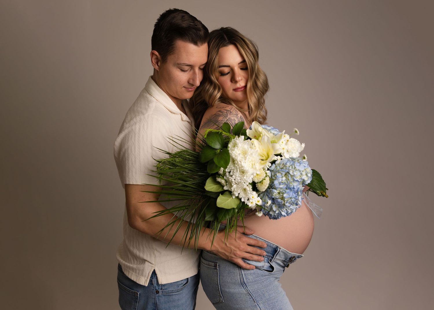 A romantic couple in casual attire embraces while holding a beautiful bouquet of white and blue hydrangeas with greenery.