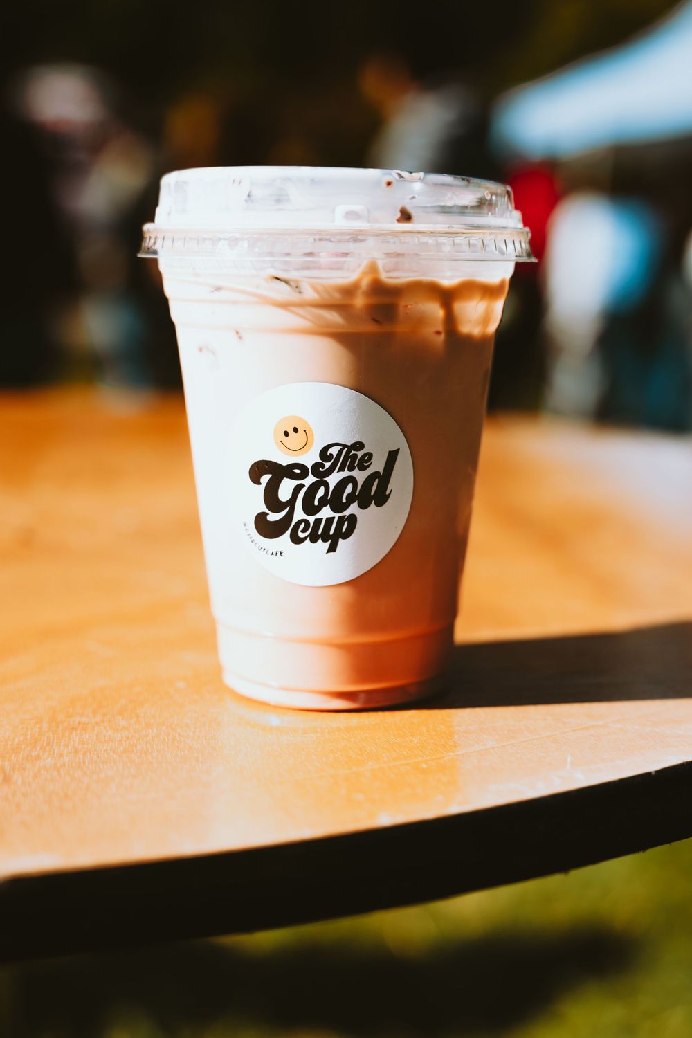 A pink iced drink in a disposable cup with 'The Good Cup' logo sits on a wooden surface in warm lighting.