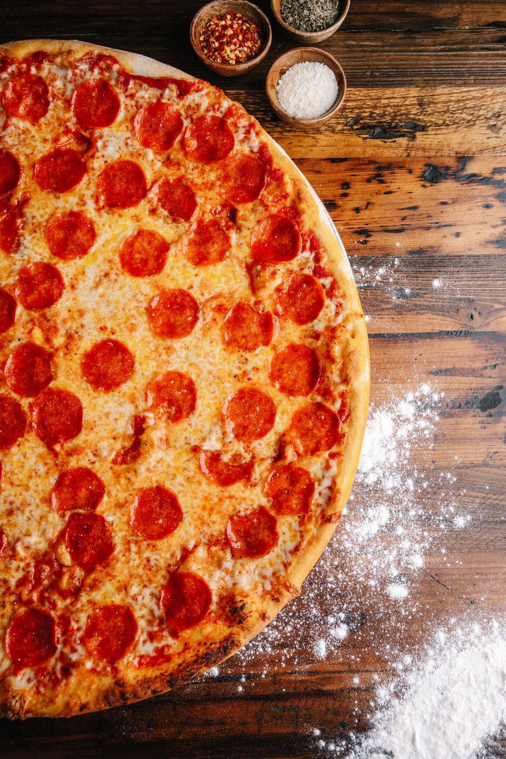Delicious pepperoni pizza with melted cheese on wooden table with scattered flour and seasoning bowls.