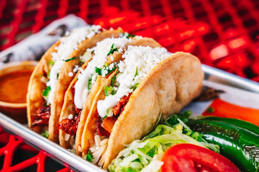 Three soft shell tacos filled with meat and garnished with fresh vegetables on a metal serving tray with red background.