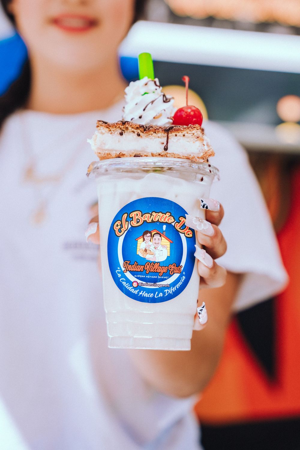 A milkshake topped with whipped cream, cherry and chocolate drizzle in a branded blue logo cup being held up.