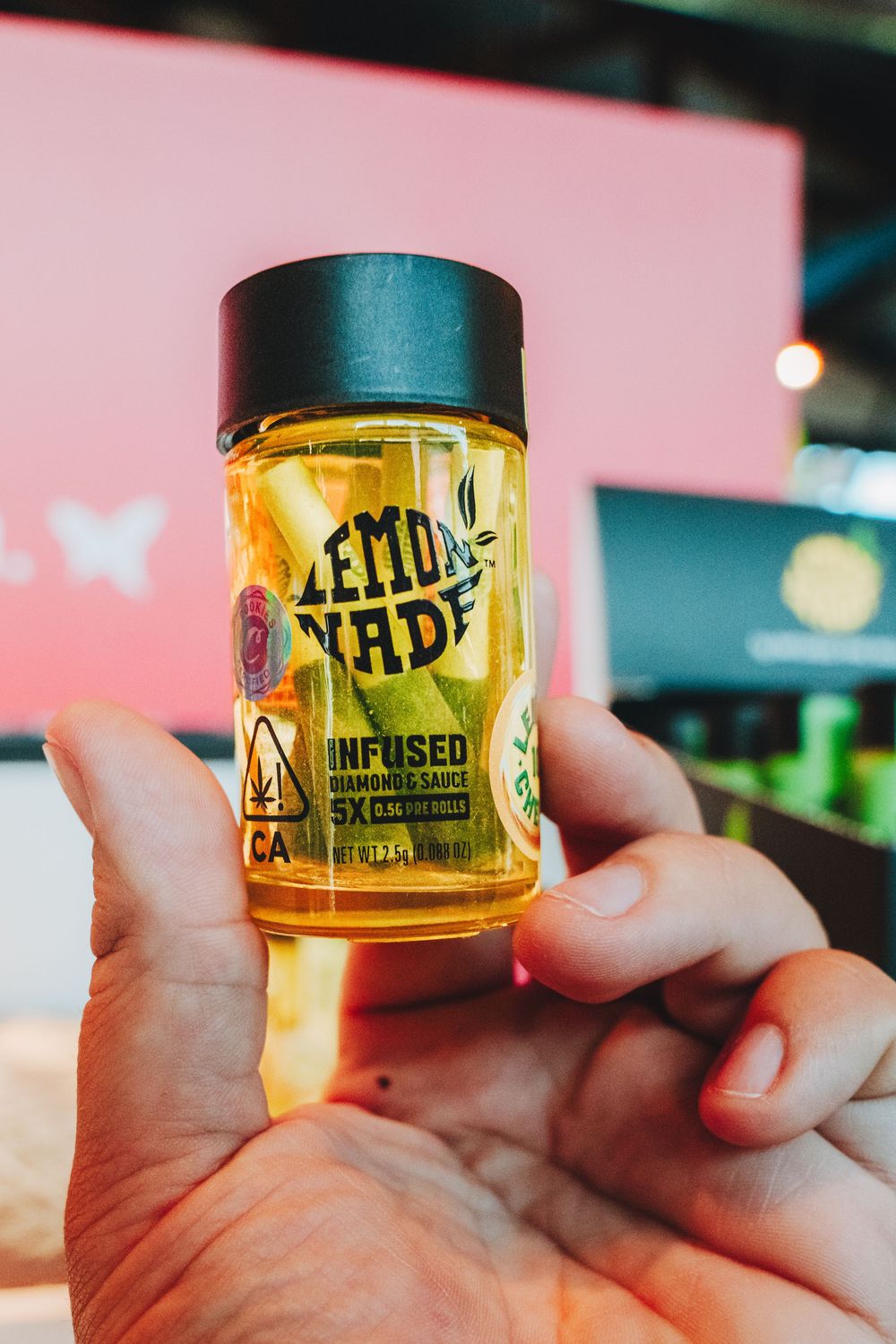 Hand holding a small glass jar of yellow Lemon Fade infused cannabis product against a pink background.