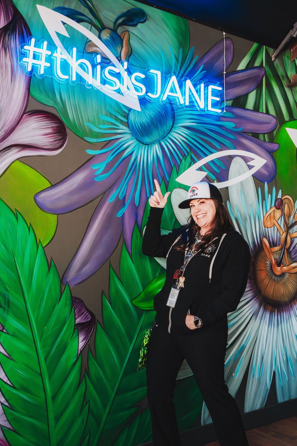 A person stands against a vibrant floral mural wall with neon blue lighting and tropical leaves in the background.