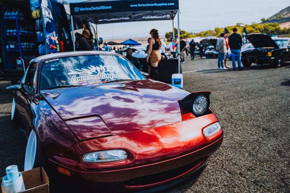 Red Mazda Miata at a car show with visitors looking at the classic sports car during an outdoor event.