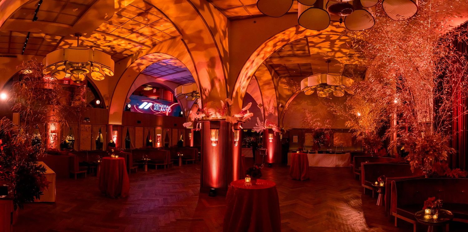 A grand ballroom venue with warm amber lighting and elegant chandeliers illuminates multiple round dining tables.