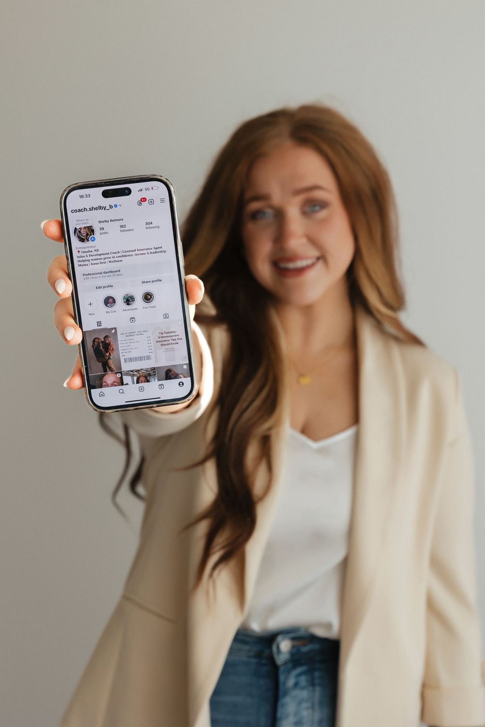 Person in beige blazer and white top holds up iPhone displaying social media profile while posing against neutral wall.