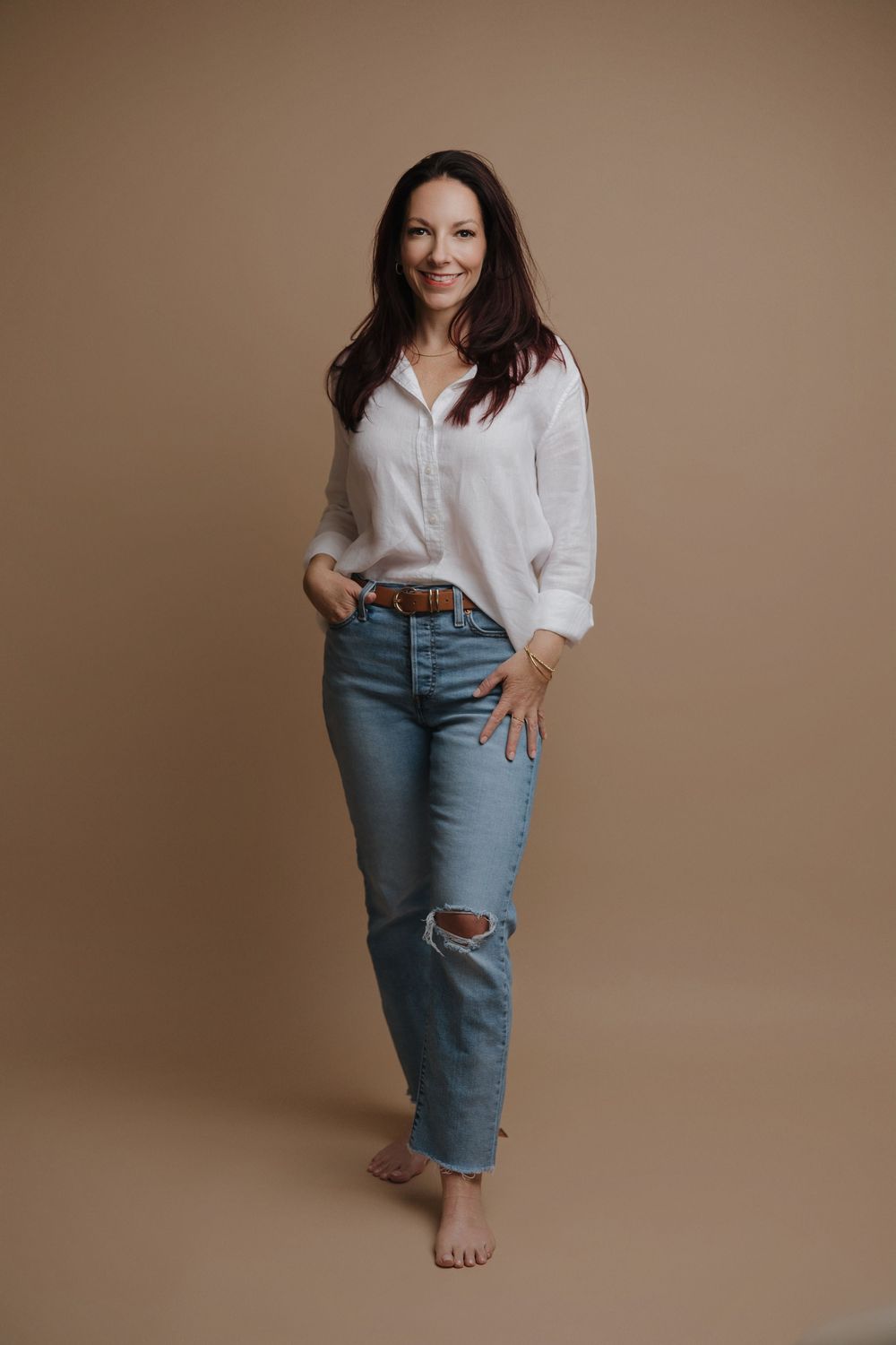 Woman in white button-up shirt, ripped jeans, and brown belt poses barefoot against a beige background.