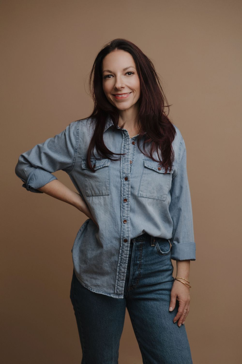 A smiling brunette woman poses confidently in a denim shirt and jeans against a warm beige background.