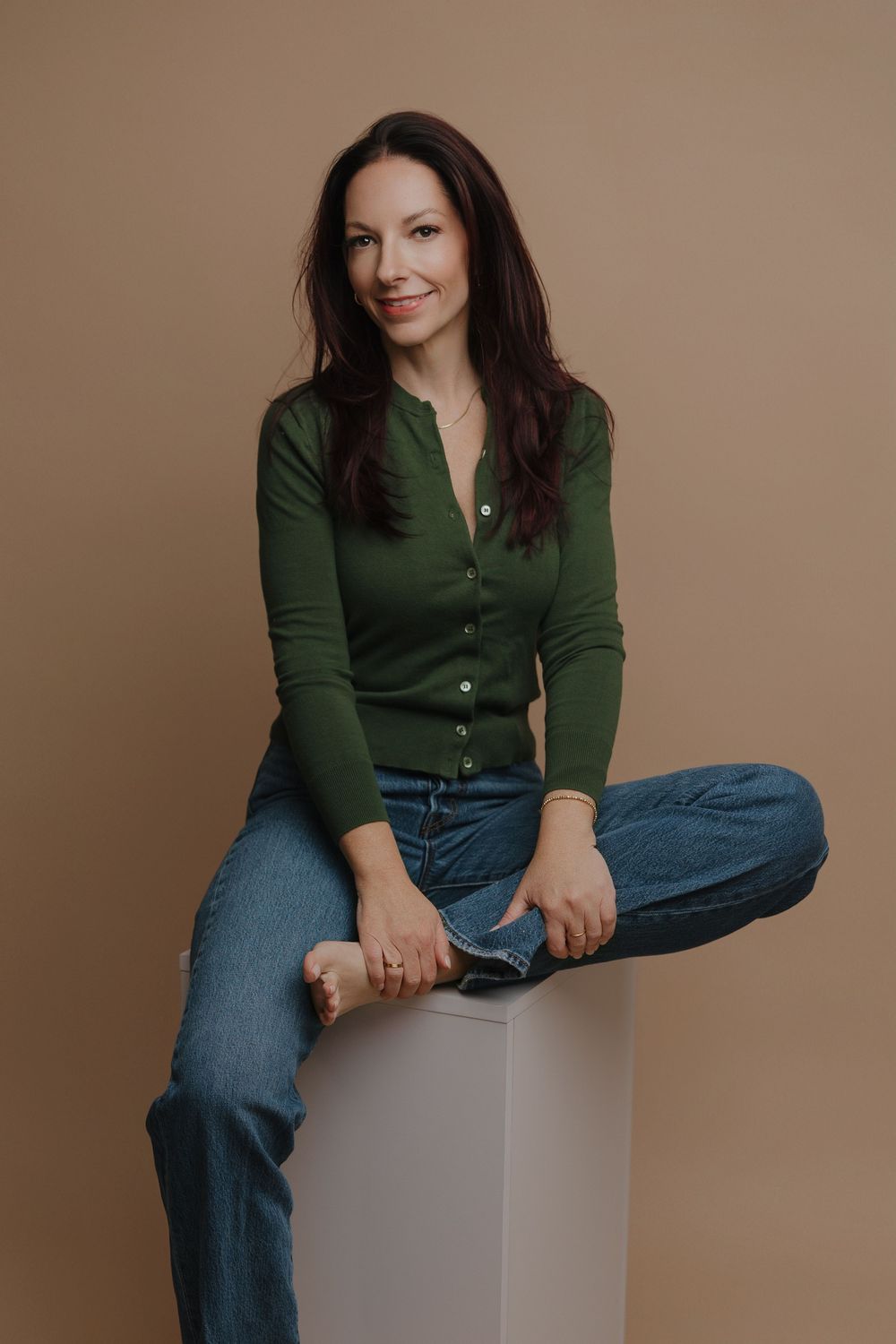 Woman in green cardigan and jeans sitting cross-legged on white cube against beige background, smiling naturally.