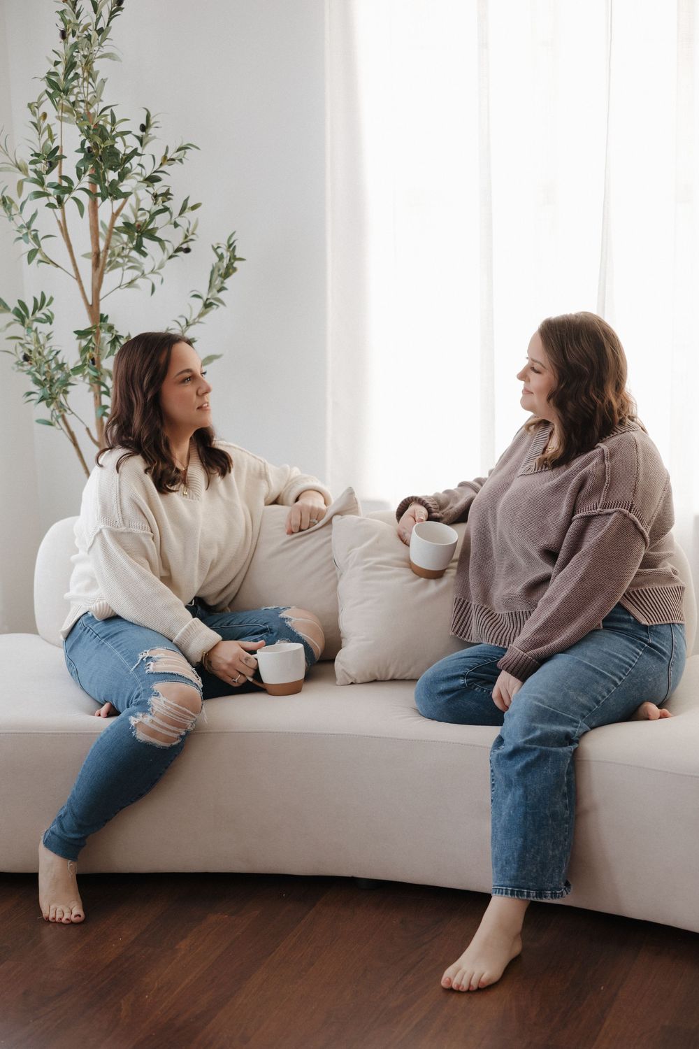 Two women sitting on a white couch, holding mugs and having a conversation in a bright, cozy living room.