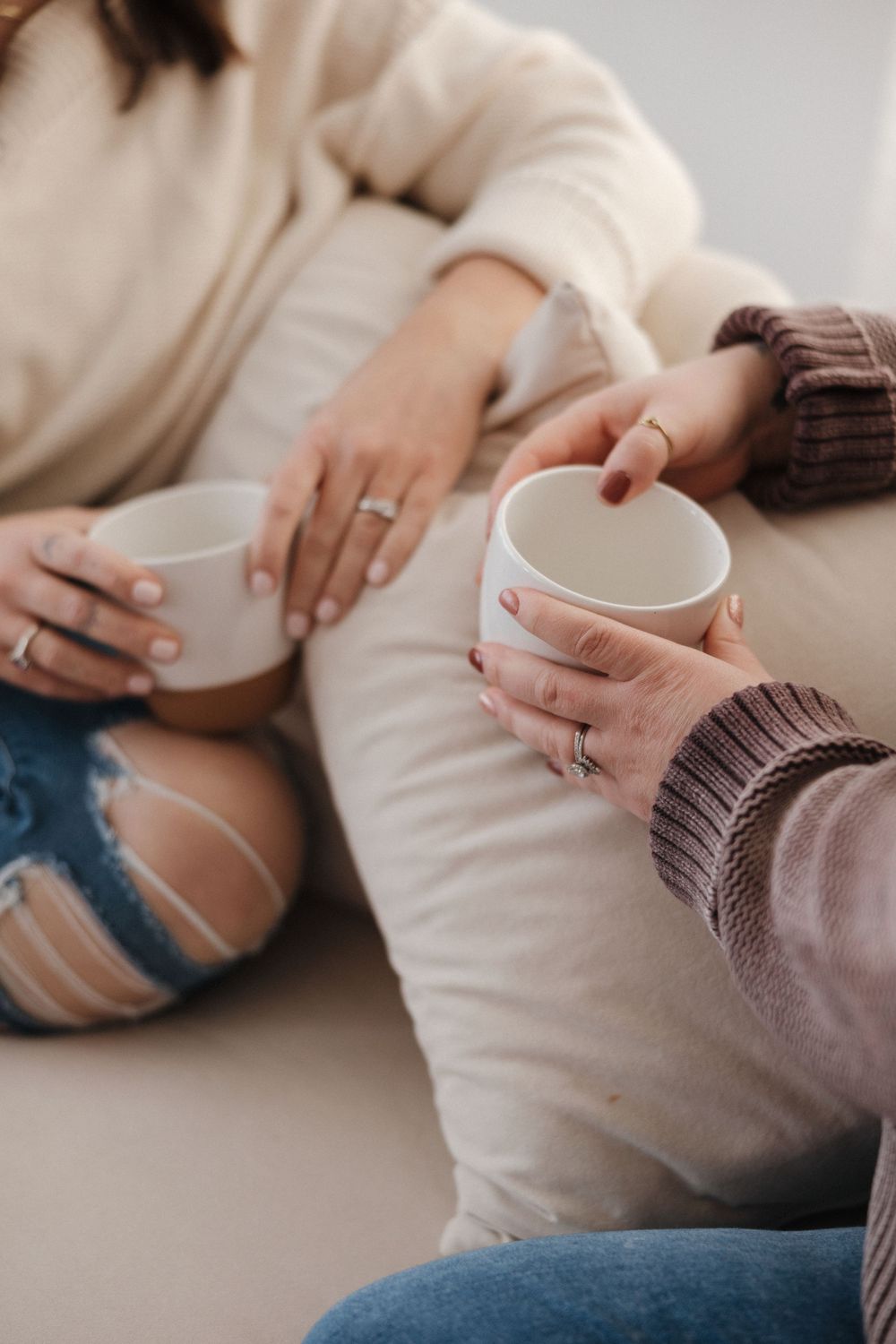 Two women sitting together on a couch, each holding a white ceramic mug, wearing cozy sweaters and jeans.