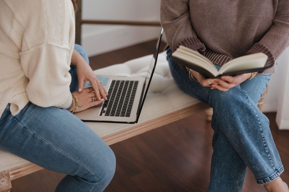 Two people sitting on a bench, one using a laptop and the other reading a book, in a cozy indoor setting.