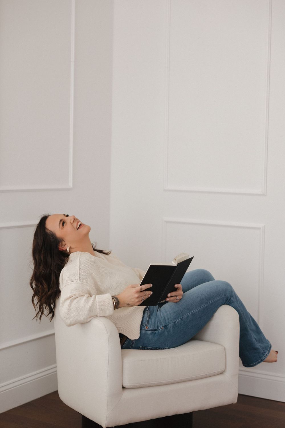 A woman relaxes in a white armchair, holding a black book and looking up thoughtfully, wearing jeans and a cream sweater.