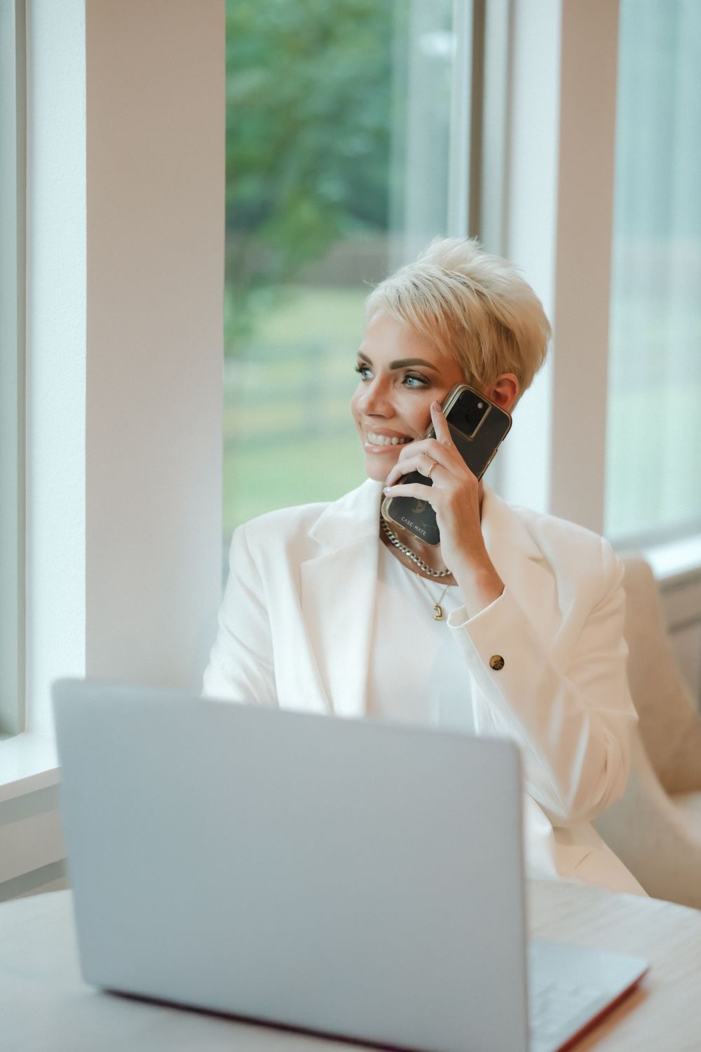Professional in a white blazer works at a laptop while having a business conversation on a mobile phone.