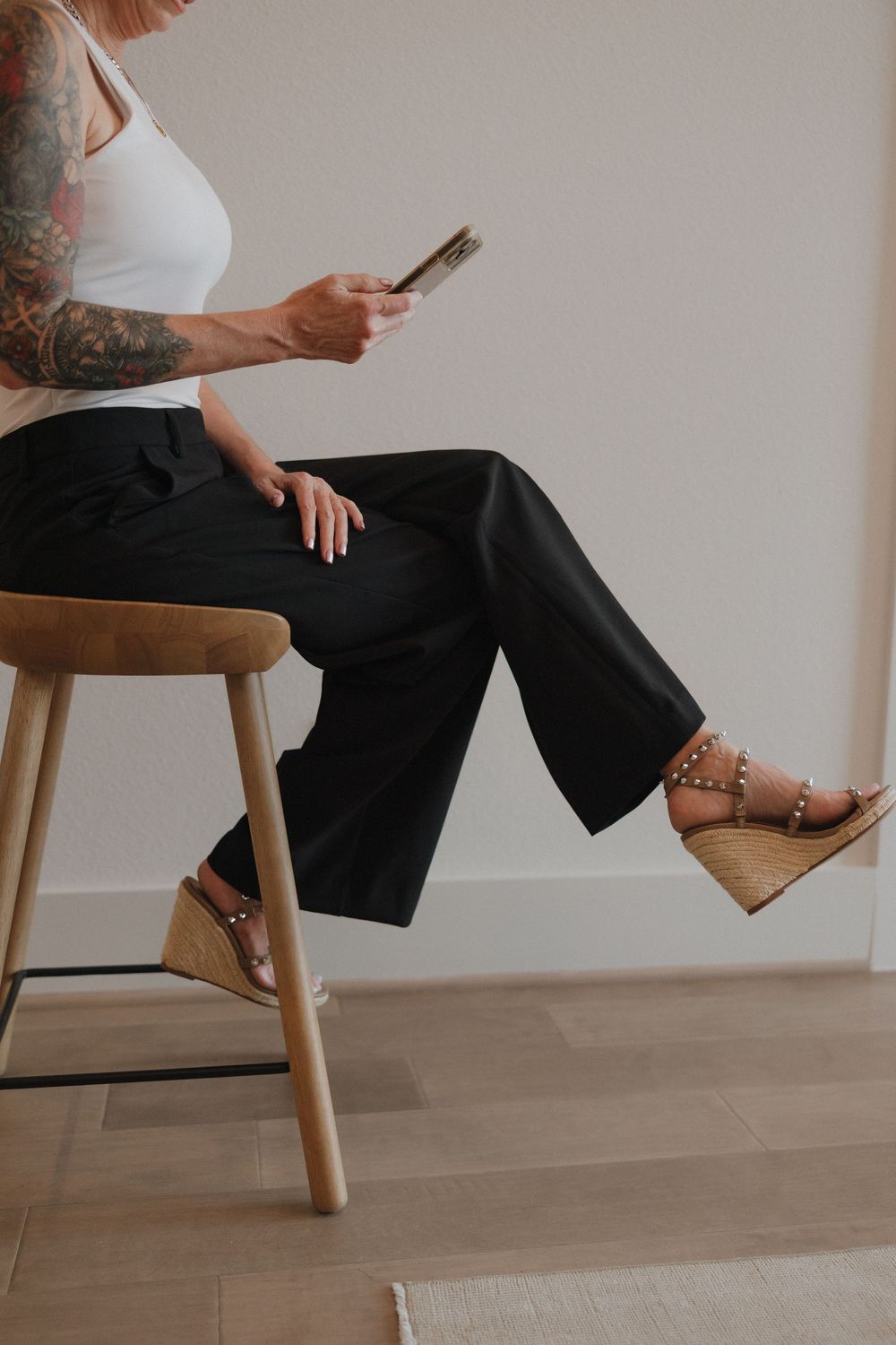 Someone wearing black wide-leg pants and cork wedge sandals sits on a wooden stool while checking their phone.