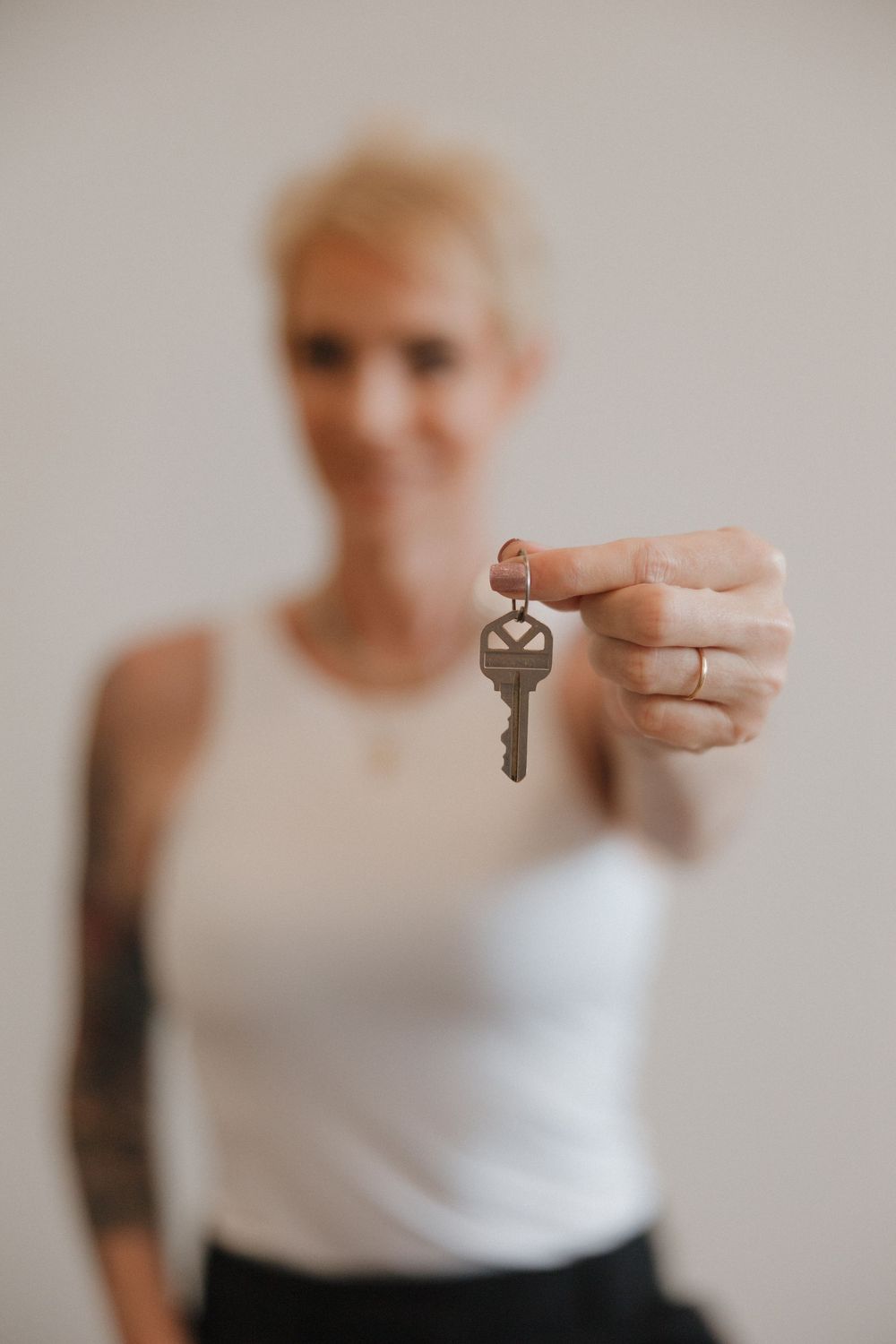 Hand holding silver key with blurred figure wearing white tank top and tattoos in background.