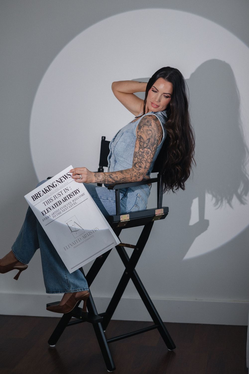 A seated figure in denim clothing holds a newspaper while posing against a white wall with dramatic shadow angles.